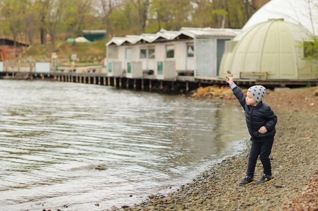 Ein kleiner Junge mit grauem Hut und dunkelblauer Jacke geht am Meer spazieren und wirft Steine darauf Karte mit selektivem Fokus