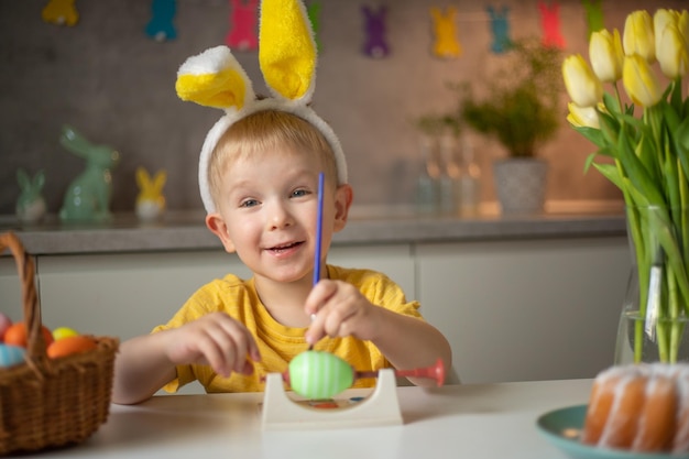 Ein kleiner Junge mit Hasenohren bereitet bemalte Ostereier für Ostern vor, während er am Küchentisch sitzt Frohe Ostern