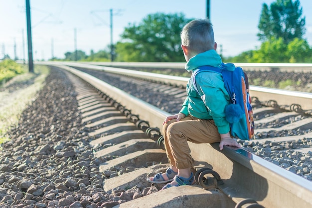 Ein kleiner Junge mit Rucksack sitzt auf den Schienen Ruhen Sie sich während eines Spaziergangs aus Warten auf den Zug