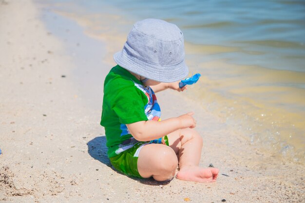 Ein kleiner Junge sitzt mit Panamahut auf dem Sand am Wasserrand und spielt mit einem Schaufelh...