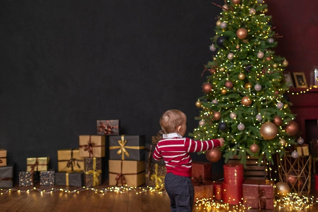 Ein kleiner Junge steht vor einem Weihnachtsbaum und Kisten mit Geschenken, die an einer schwarzen Wand stehen