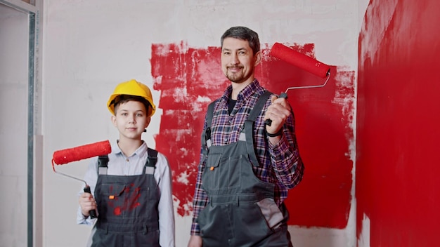 Ein kleiner Junge und sein lächelnder Vater stehen im Raum und halten mit roter Farbe bedeckte Walzen