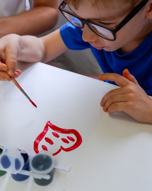Ein kleiner Junge zeichnet mit Pinsel und Farbe ein rotes Herz auf weißes Papier