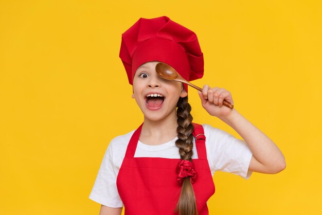 Ein kleiner Mädchenkoch spielt mit hölzernen Löffeln Kochen für Kinder Ein Kind in einer roten Schürze und in einer Kappe auf einem gelben getrennten Hintergrund