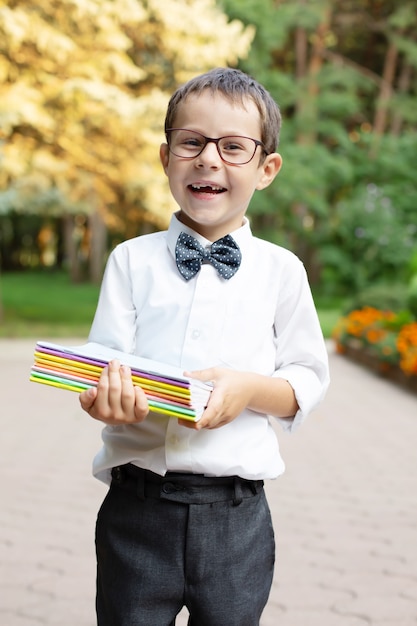 Ein kleiner schöner fröhlicher Schuljunge mit Brille in einem weißen Hemd hält Notizbücher in den Händen