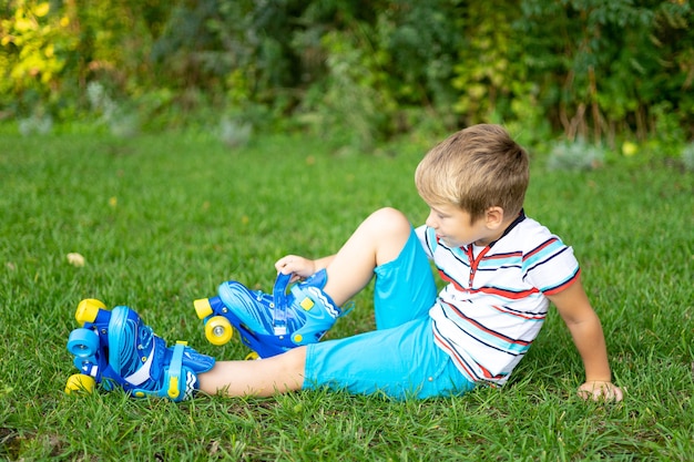 Ein kleiner süßer blonder Junge in Rollschuhen sitzt auf dem Gras lächelt und zieht Rollschuhe an