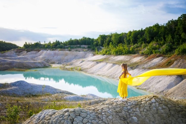 Ein kleines 67-jähriges ukrainisches Mädchen in einem gelben Kleid mit Flügeln aus gelbem Stoff am See Blauer Himmel und gelber Stoff in den Farben der ukrainischen Flagge