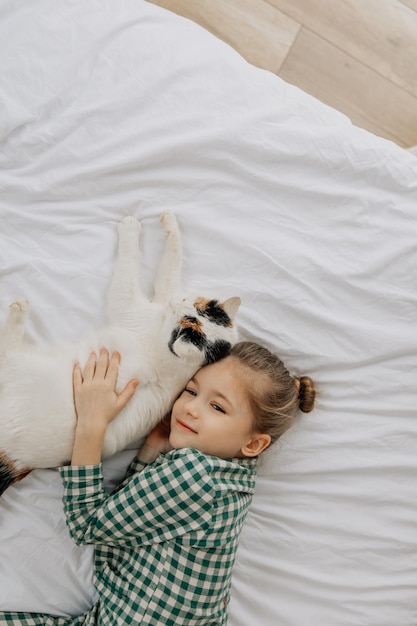 Ein kleines blondes Mädchen im Schlafanzug umarmt eine Hauskatte in ihrem Schlafzimmer das Konzept eines gemütlichen Heimmorgens