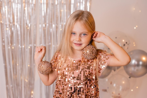 Ein kleines blondes Mädchen in einem glänzenden Kleid spielt mit Weihnachtskugeln beim Neujahrskarneval