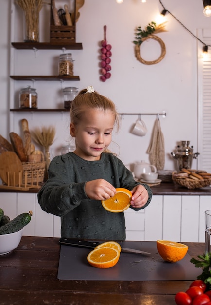 Ein kleines blondes Mädchen in einem grünen Pullover sitzt am Tisch und isst eine Orange