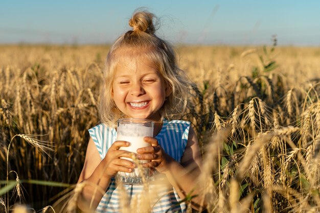 Ein kleines blondes Mädchen in einem Roggenfeld lächelt, ein glückliches Kind in den Ohren mit einem Glas Milch