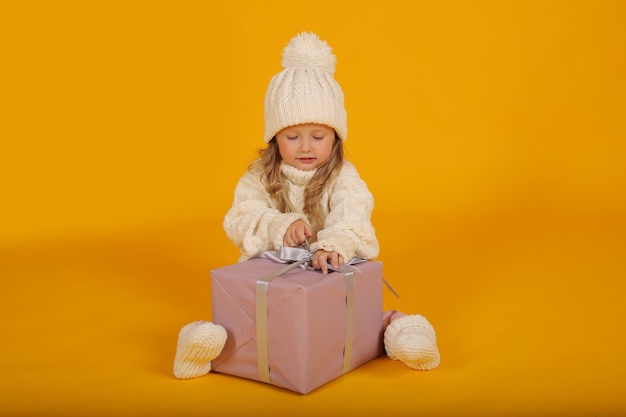ein kleines blondes Mädchen in einem weißen Strickpullover und Socken mit einer Geschenkbox in den Händen