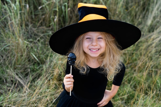 Ein kleines blondes Mädchen in schwarzem Kostüm und Hexenhut, Halloween mit einem Süßigkeitenschädel in den Händen, ein fröhliches Kind.