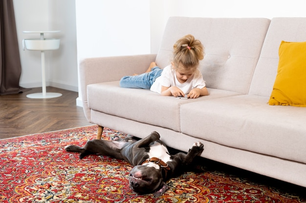 Ein kleines blondes Mädchen zu Hause auf dem Sofa mit einem Handy und einem großen Hund, Kindern und Geräten.