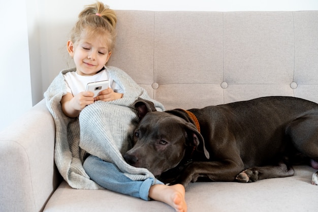 Ein kleines blondes Mädchen zu Hause auf dem Sofa mit einem Handy und einem großen Hund, Kindern und Geräten.