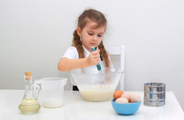 Ein kleines charmantes Mädchen in einer Schürze bereitet Teig aus Mehl, Eiern und Milch zu, Kind mischt Zutaten in Glasschüssel, kleiner Koch bereitet Essen zu. Hausgemachte Kuchen.