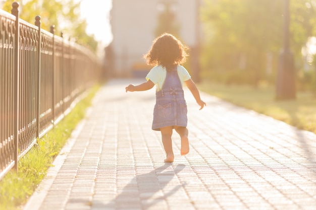 Ein kleines dunkelhäutiges Mädchen mit lockigem Haar in einem Denim-Sommerkleid geht an einem sonnigen Tag auf die Straße