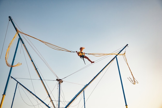 Ein kleines fröhliches Mädchen fliegt auf federnden hellen Gummibändern und springt auf ein Trampolin, um den lang erwarteten Urlaub in der warmen Sonne zu genießen