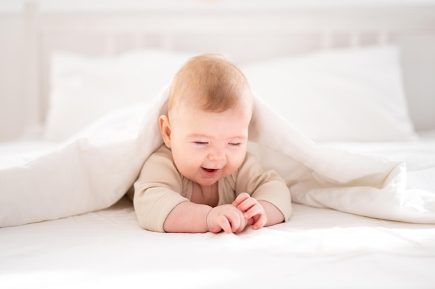 Ein kleines gesundes Baby in einem Baumwollbody liegt auf dem Bauch unter einer Decke auf einem Bett auf weißem Bettzeug im Schlafzimmer zu Hause und schaut auf die Kamera lächelt