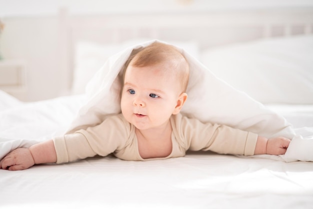 Ein kleines gesundes Baby in einem Baumwollbody liegt auf dem Bauch unter einer Decke auf einem Bett auf weißem Bettzeug im Schlafzimmer zu Hause und schaut auf die Kamera lächelt