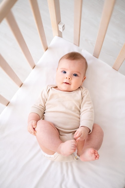 Ein kleines gesundes Baby in einem Baumwollbody liegt auf dem Rücken auf einem Kinderbett auf weißer Bettwäsche im Schlafzimmer des Hauses und schaut auf die Kamera lächelt