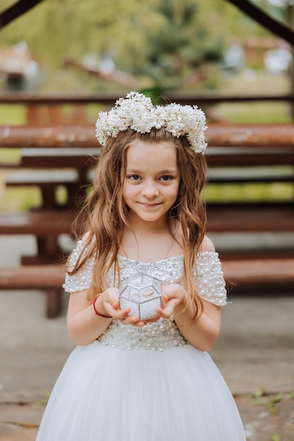 ein kleines glänzendes Mädchen mit Blumen auf dem Kopf in einem weißen Kleid, das Hochzeitsringe für die Hochzeitszeremonie hält