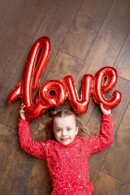 Ein kleines glückliches Mädchen in rotem Pyjama mit einem großen Ballon mit der Aufschrift Love liegt auf einem dunkelbraunen Holzhintergrund auf dem Boden und lacht über das Konzept des Valentinstags, einen Ort für Text