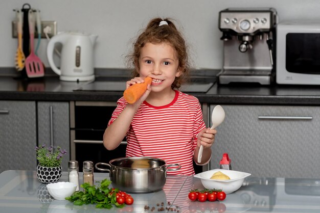 Ein kleines hübsches Mädchen kocht und isst Karotten in der Küche aus frischem Gemüse