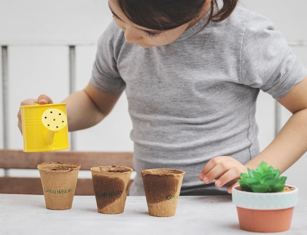 Ein kleines kaukasisches Mädchen mit grauem Rollkragen hält eine gelbe Gießkanne mit ihren Händen und wässert Pappgläser