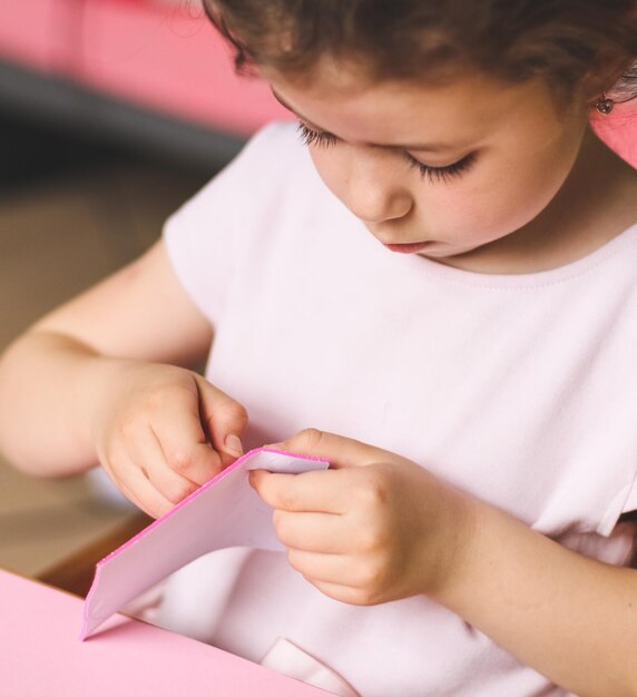 Ein kleines kaukasisches Mädchen zieht mit den Fingern einen rosafarbenen Filzaufkleber ab, während es an einem Kindertisch sitzt