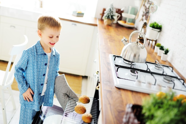 Ein kleines Kind, ein Junge im blauen Pyjama mit einem Handschuh an der Hand, backt Brötchen und lächelt vor dem Hintergrund einer modernen Küche