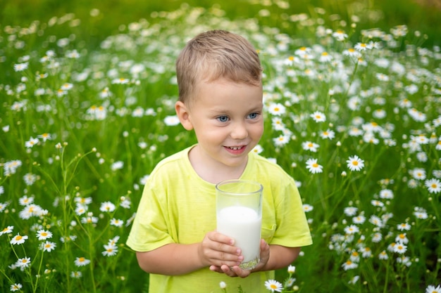Ein kleines Kind, ein Junge in Kleidung aus natürlicher Baumwolle, geht um das Feld herum und hält ein Glas Milch in den Händen, genießt das Leben, genießt die Natur