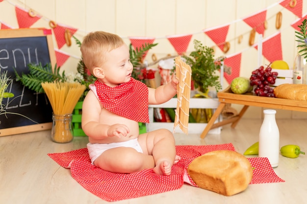 Ein kleines Kind, ein sechs Monate alter Junge, sitzt mit einem Laib Brot auf dem Küchenboden und hält eine Sushi-Matte in der Hand