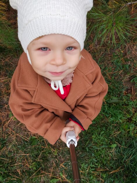 Ein kleines Kind in einem Herbstmantel lächelt in die Kamera und nimmt einen Selfie-Stick Mädchen im Park Herbst