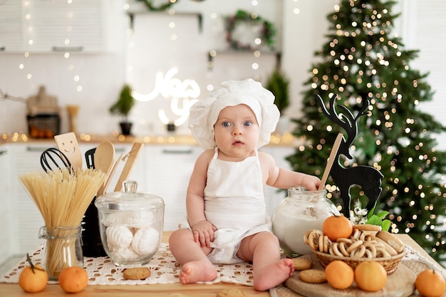 ein kleines Kind in einem Kochkostüm mit Schürze sitzt auf dem Tisch und hilft, ein festliches Abendessen vorzubereiten vor dem Hintergrund eines Weihnachtsbaums und eines Neujahrsdekors, das sich auf den Urlaub vorbereitet