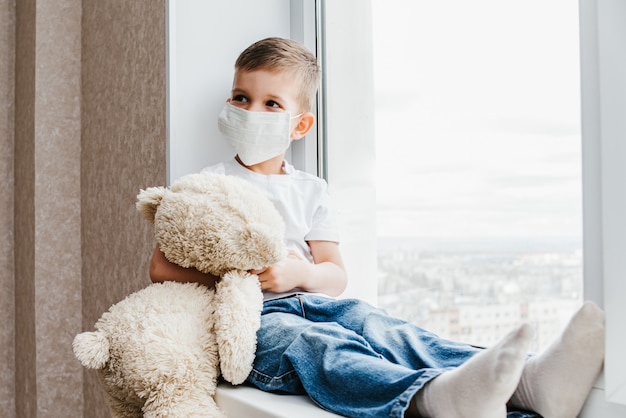 Ein kleines Kind in einer Maske sitzt zu Hause in Quarantäne und schaut an einem Ort mit einem Teddybär aus dem Fenster. Prävention von Coronavirus und Covid - 19. Konzept