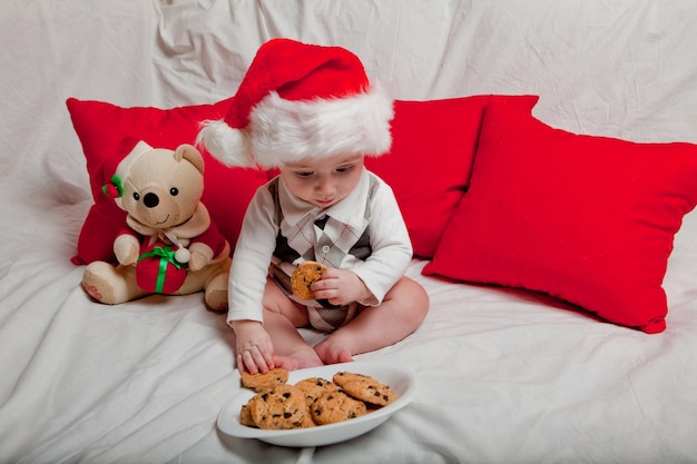 Ein kleines Kind in einer roten Mütze isst Kekse und Milch Weihnachtsfotografie eines Babys in einer ruten Mütze Neujahrsferien und Weihnachten