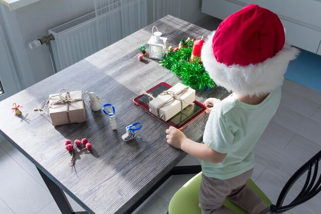 Ein kleines Kind in einer Weihnachtsmannmütze mit Weihnachtsgeschenken sitzt mit einem Tablet in den Händen an einem Neujahrstisch