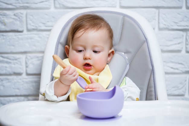 Ein kleines Kind sitzt auf einem Hochstuhl und isst mit einem Löffel Essen von einem Teller. Baby-Silikon-Utensilien zum Füttern von Babys