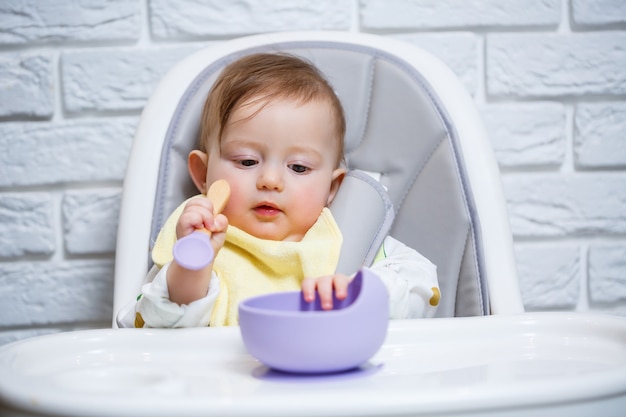 Ein kleines Kind sitzt auf einem Hochstuhl und isst mit einem Löffel Essen von einem Teller. Baby-Silikon-Utensilien zum Füttern von Babys