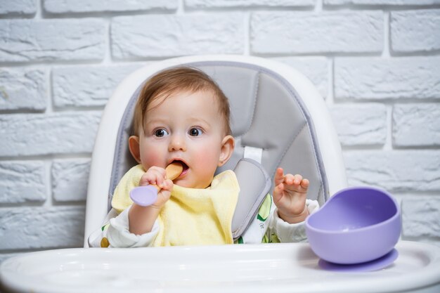 Ein kleines Kind sitzt auf einem Hochstuhl und isst mit einem Löffel Essen von einem Teller. Baby-Silikon-Utensilien zum Füttern von Babys