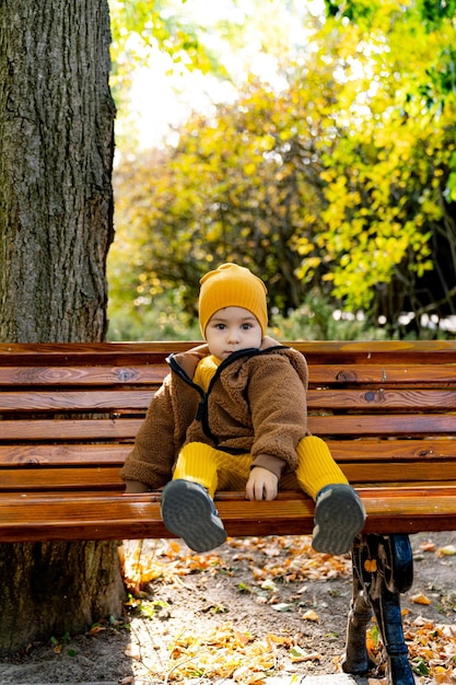 Ein kleines Kind sitzt auf einer Holzbank in einem Park. Das Kind trägt einen gelben Hut und eine braune Jacke.