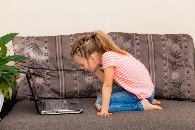 Ein kleines Kind surft auf einem grauen Sofa im Internet. Mädchen mit Laptop auf der Couch sitzend