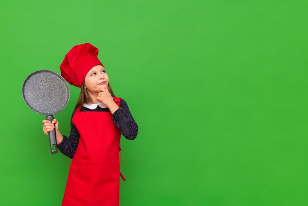 Ein kleines Kochmädchen in einem roten Hut und einer Schürze hält eine Pfannkuchenpfanne und sieht sich Ihre Werbung auf einem grün isolierten Hintergrund an Auswahl eines Berufs für einen Studenten Kopierbereich