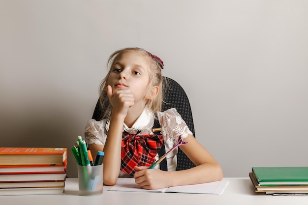 Ein kleines linkshändiges Mädchen in Schuluniform sitzt mit Büchern und Stiften an einem Tisch und überlegt, was es in sein Heft schreiben soll