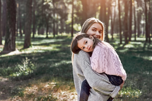 Ein kleines lockiges Mädchen im Pullover umarmt ihre Mutter im Wald. Kalte Jahreszeit, helle Sonne wird durch die Bäume gesehen. Sie trägt einen rosa Pullover und eine junge Frau trägt einen grauen Pullover