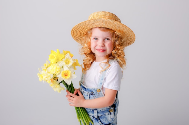 ein kleines lockiges mädchen in einem strohhut hält einen blumenstrauß aus frühlingsgelben narzissenblumen auf einem whi