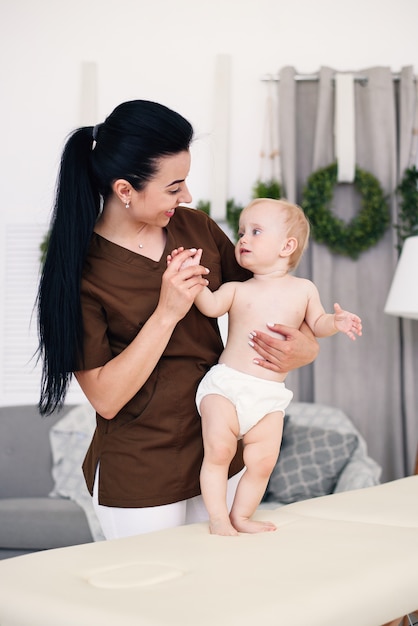 Ein kleines lustiges Baby, das Massage mit professioneller Masseurin hat. Kindermassage auf der Couch in einem modernen gemütlichen Raum. Freundlicher und freundlicher Kinderarzt.