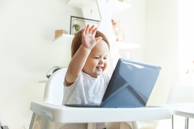 Ein kleines lustiges Baby winkt beim Laptopbildschirm, während es zu Hause am Tisch des Kindes sitzt