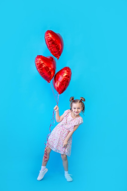 Ein kleines Mädchen auf einem blau isolierten Studiohintergrund mit roten Luftballons in Form eines Herzens hat Spaß und albert herum Platz zum Kopieren von Text Valentinstag Konzept